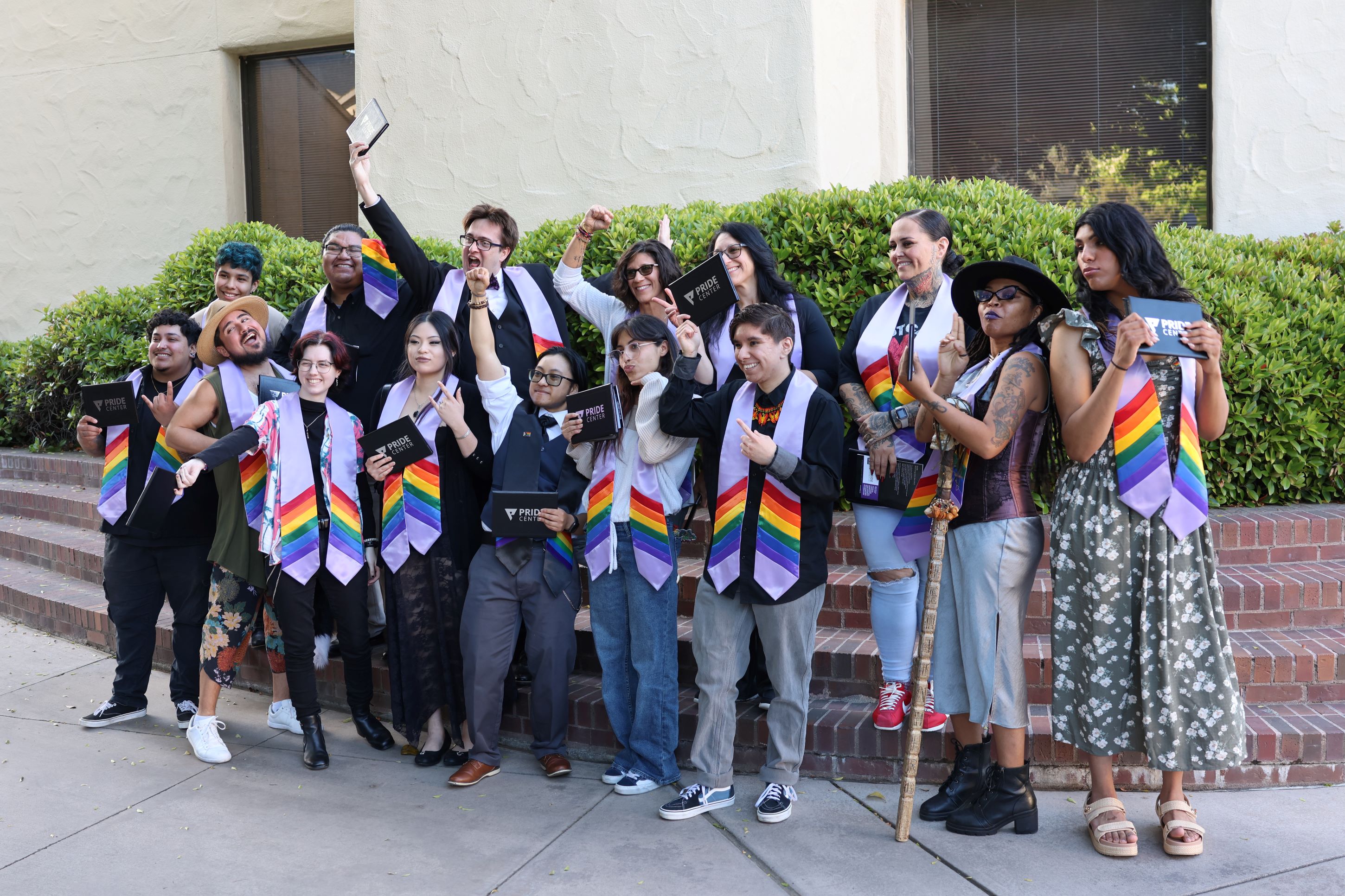 A group of students celebrate together outdoors during a Lavender Ceremony, wearing rainbow stoles and holding Pride Center programs.