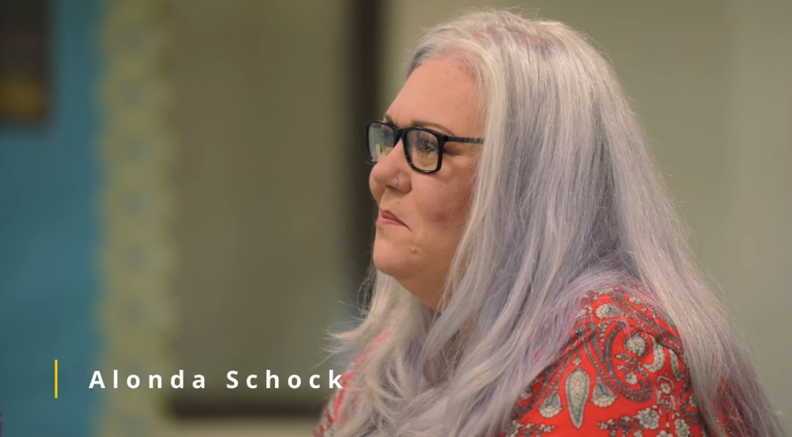 Video thumbnail showing a person with long light gray hair and glasses, seated and listening during a conversation.