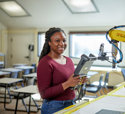 A black female student stands smiling in front of a robot arm in a bright modern classroom