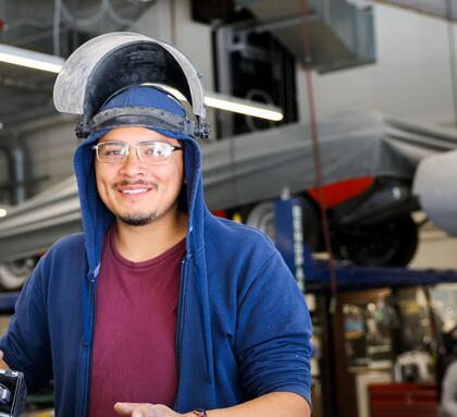 An Auto Body student wearing safety glasses and a raised welding helmet smiles while working on a vehicle in an automotive lab.