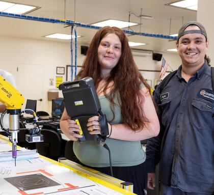 Two Robotics & Automation Technology students stand beside an industrial robotic arm, with one holding a handheld programming device in a robotics lab.