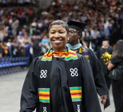 A Delta College graduate wearing a cap, gown, and colorful stole smiles while walking during the commencement ceremony, with graduates and guests in the background.