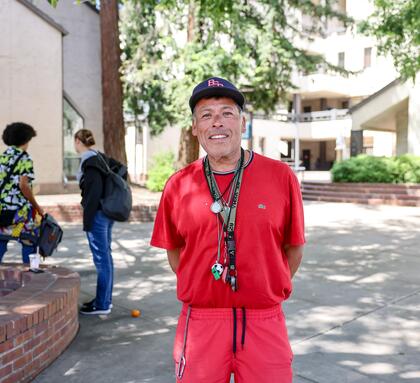 An older student wearing a red shirt and baseball cap stands smiling on a shaded campus walkway with other students in the background.
