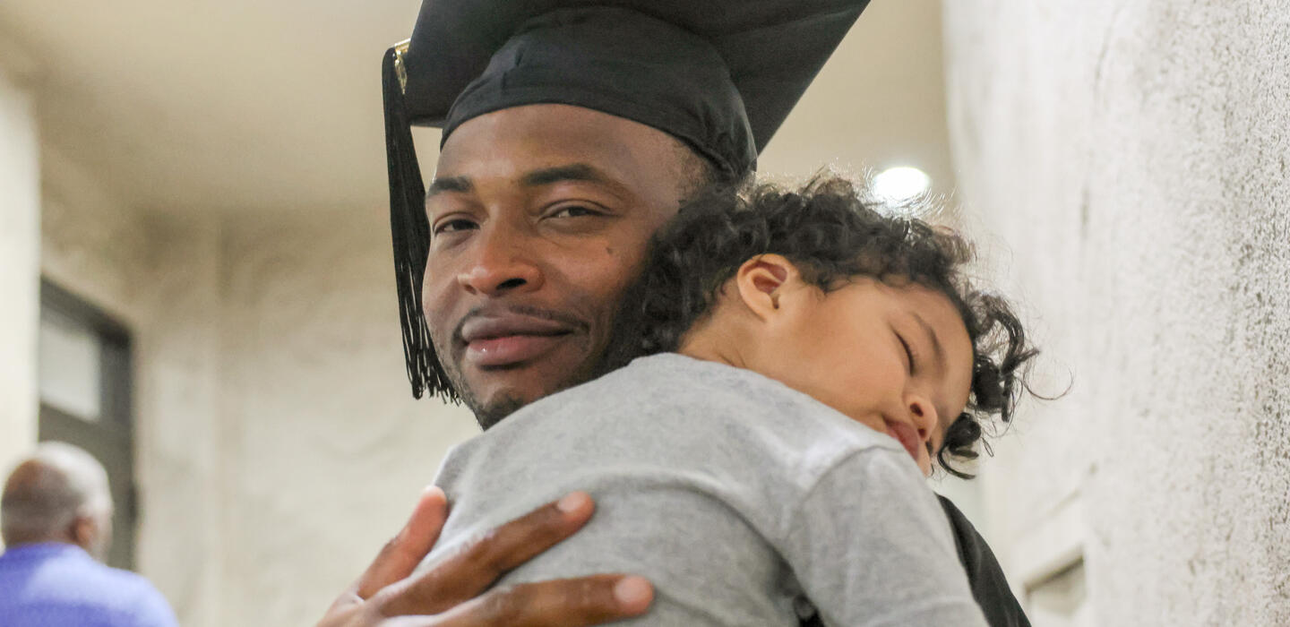A student celebrates graduation while holding his sleeping son