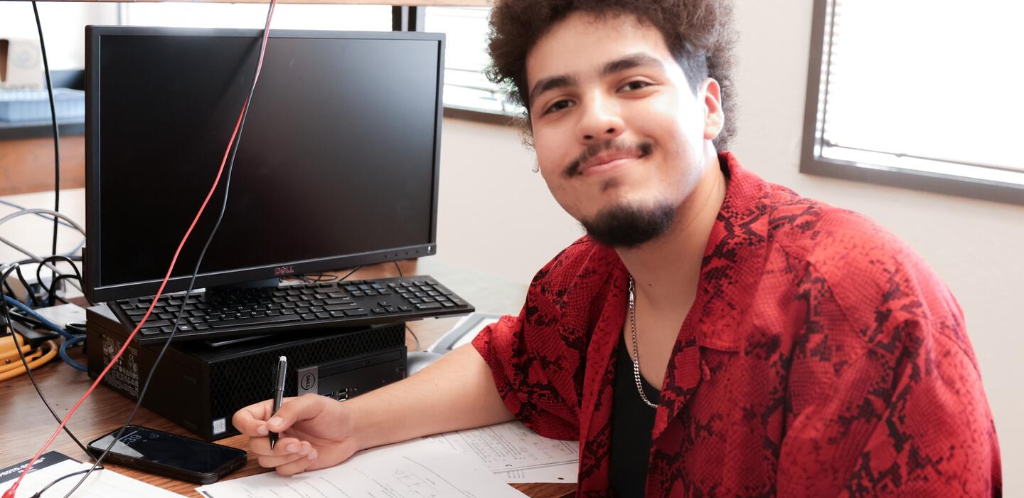 A student sits at his work station and smiles for the camera.