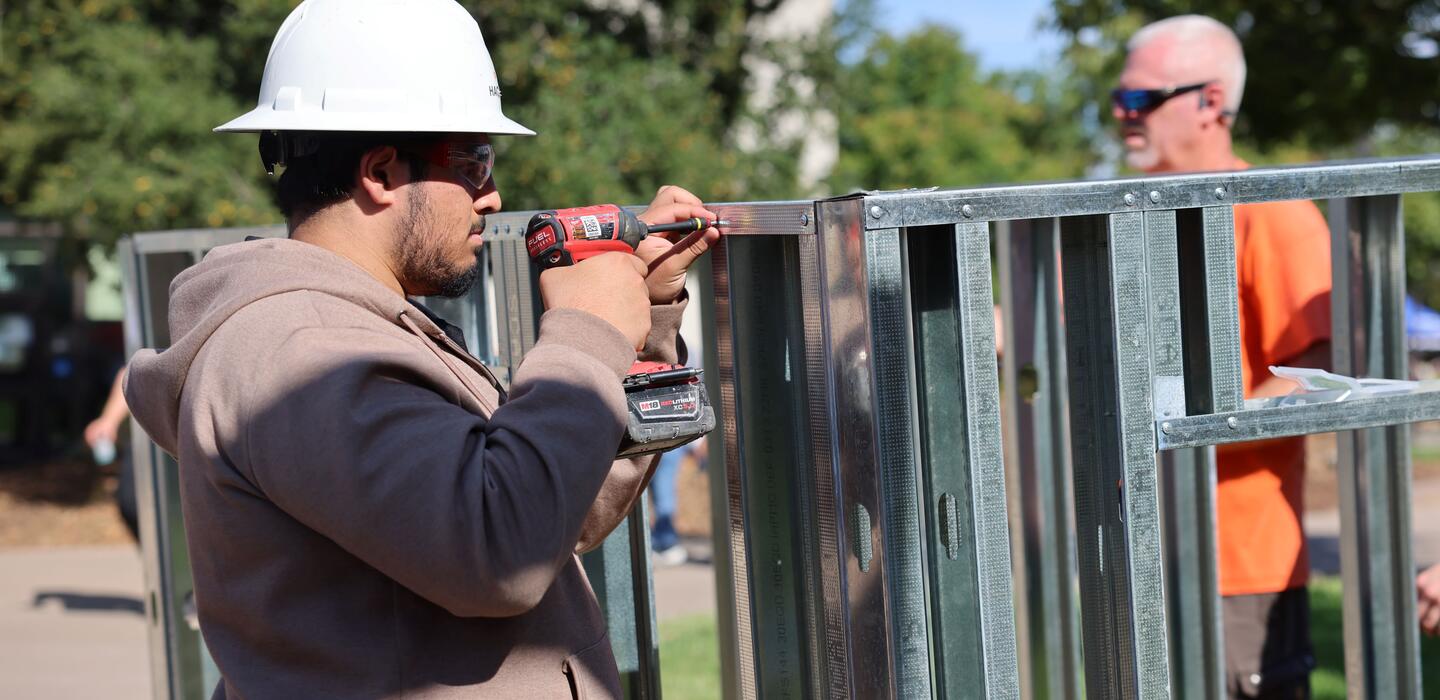Student wearing a hard hat uses a power drill to fasten metal framing