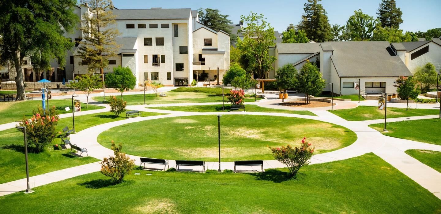 View of the San Joaquin Delta College campus featuring landscaped lawns, curved pedestrian walkways, benches, mature trees, and campus buildings surrounding a central green space.