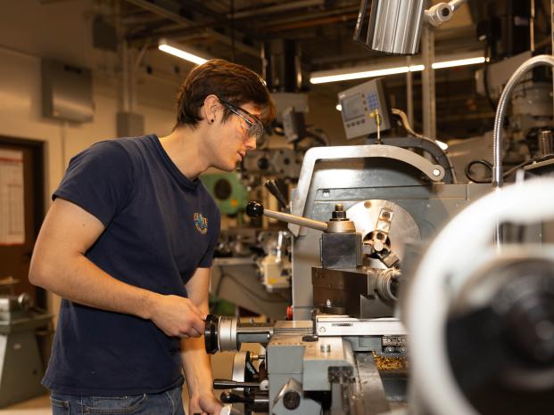 A student focused on operating a machine in a workshop setting, wearing safety gear and surrounded by heavy machine.