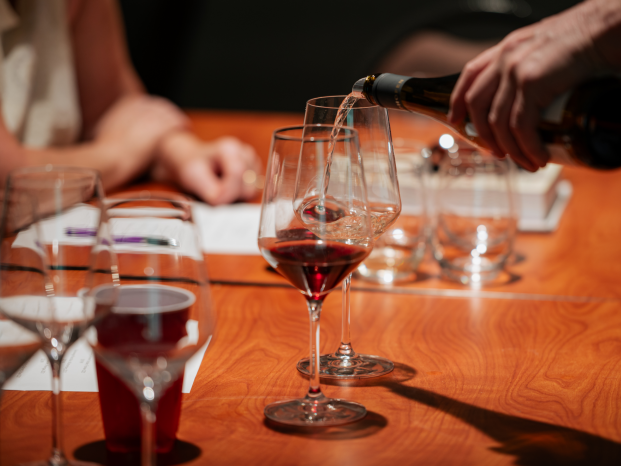 A person pouring red wine from a bottle into a clear glass on a wooden table.