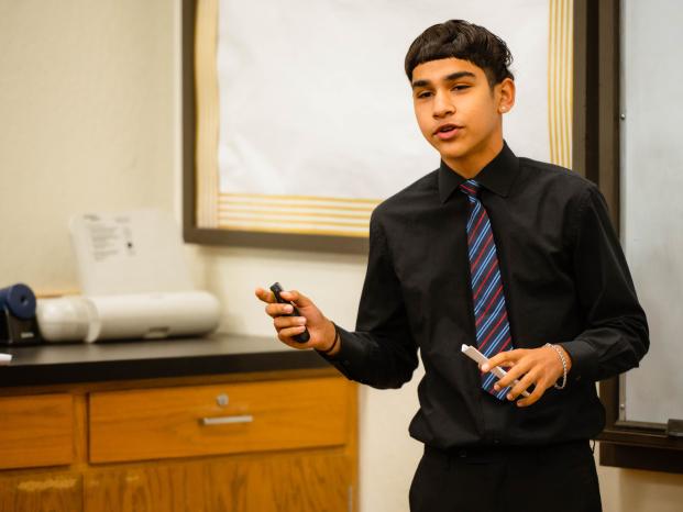A young man in a black shirt and tie stands confidently in front of a whiteboard, ready to present.