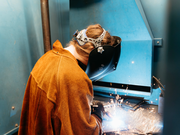 A female student practices metal welding in proper protective gear and factory setting