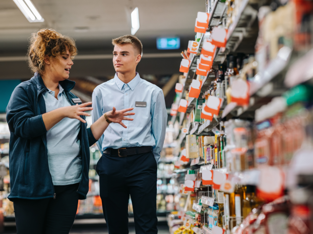 A female retail clerk guides a young male novice through a supermarket aisle, both engaged in conversation.