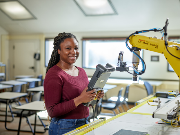 A black female student stands smiling in front of a robot arm in a bright modern classroom