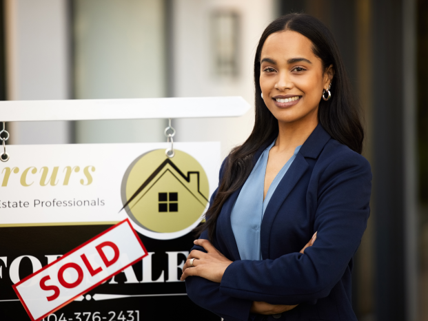 A smiling real estate agent stands next to a "Sold" sign in front of a house, celebrating a successful sale.