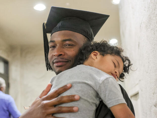 A student celebrates graduation while holding his sleeping son