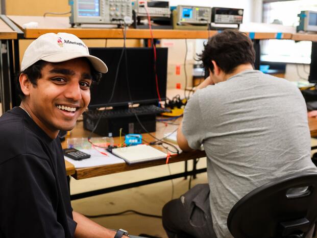 A student smiles at the camera while working on an electronics project in a lab, seated next to another student.