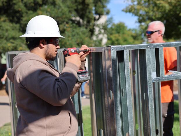 Student wearing a hard hat uses a power drill to fasten metal framing