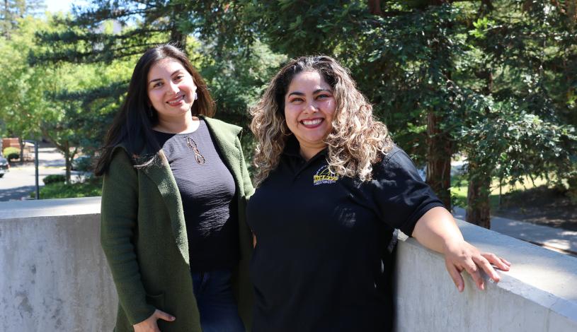 Two people standing next to each other smiling with tall green trees in the background