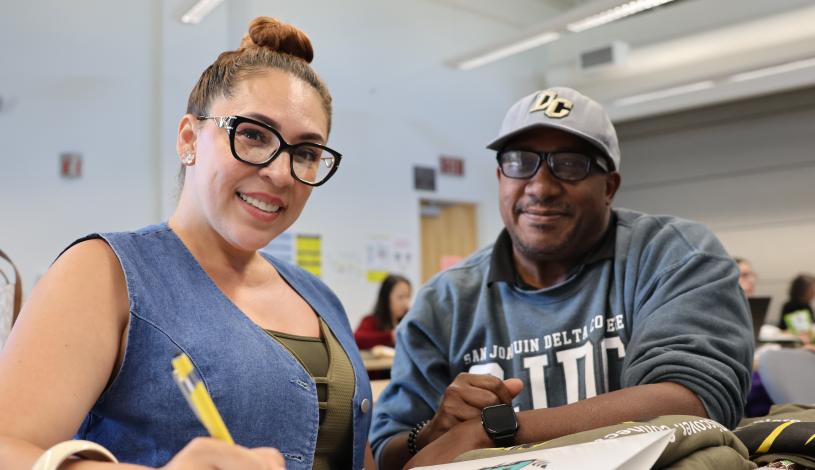 Two people sitting at a desk one holding a pen with a folder and both smiling