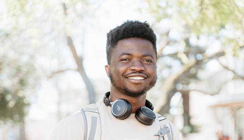 Student standing in the quad center of campus with hands in pockets smiling and wearing headphones and backpack