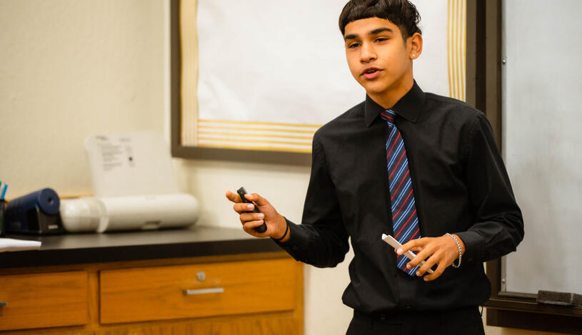 A young man in a black shirt and tie stands confidently in front of a whiteboard, ready to present.