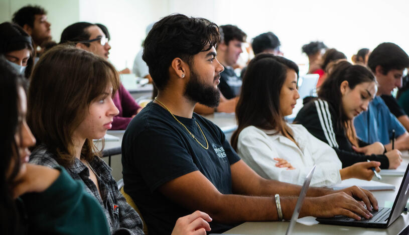 Students in a lecture hall attentively using laptops to take notes and engage with the lecture content.