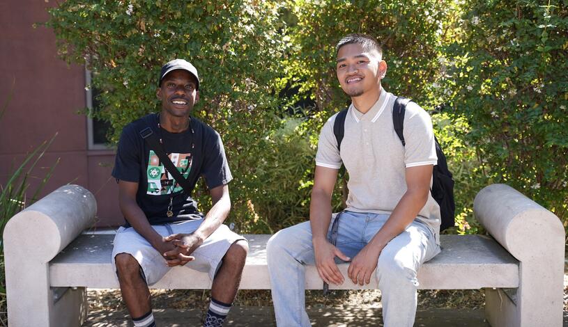 Two students sit on a bench and pose for a photo