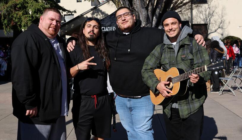 Four students stand together outdoors on campus, smiling for the photo. One of them holds an acoustic guitar.