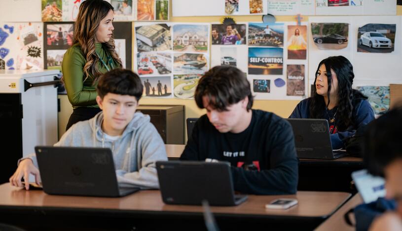 Two students are sitting down at their classroom desk looking at their laptops while a teacher is talking to another student behind them.