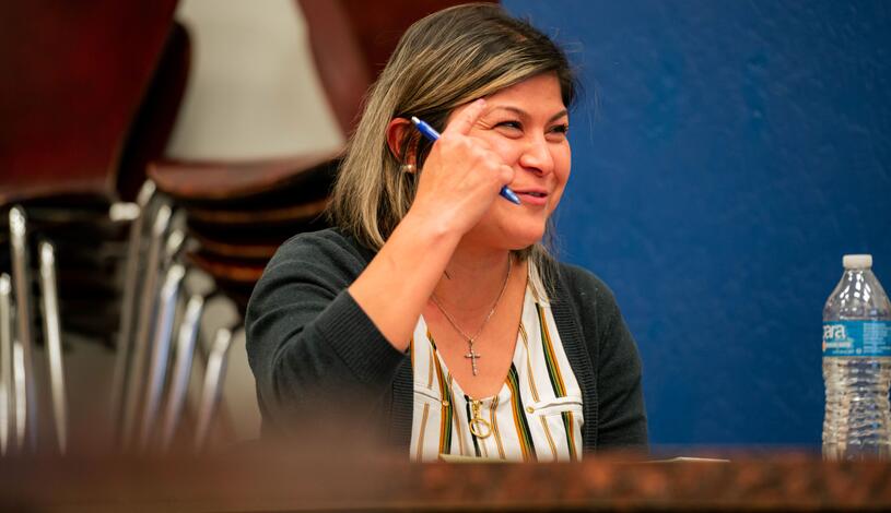 A smiling student in an English class points to her head with her finger, suggesting she has an idea during class.
