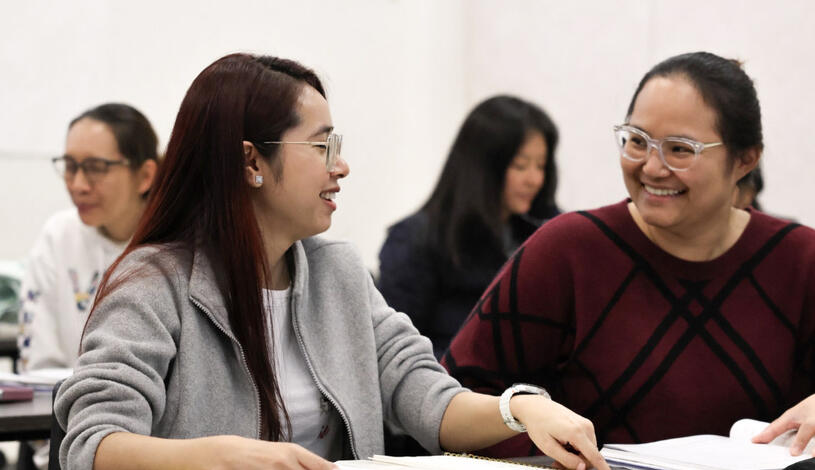 Two students sit at a classroom desk smiling and talking while looking at an open notebook, with other students studying in the background.