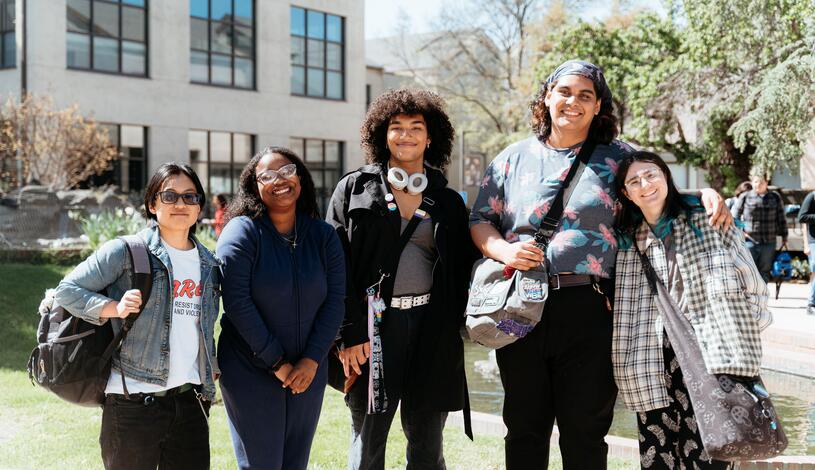 Five students stand together smiling outdoors on the Delta College campus near a pond, with campus buildings and trees in the background.