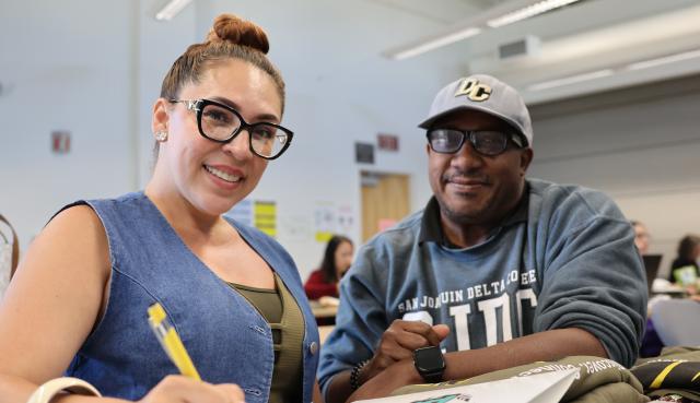 Two people sitting at a desk one holding a pen with a folder and both smiling