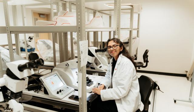 Electron Microscopy student sitting at desk with lab equipment wearing a lab coat and smiling softly