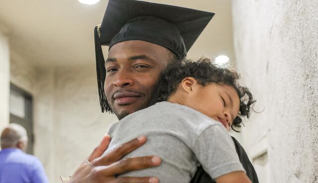 Photo of an African American graduate holding his young child after the ceremony.