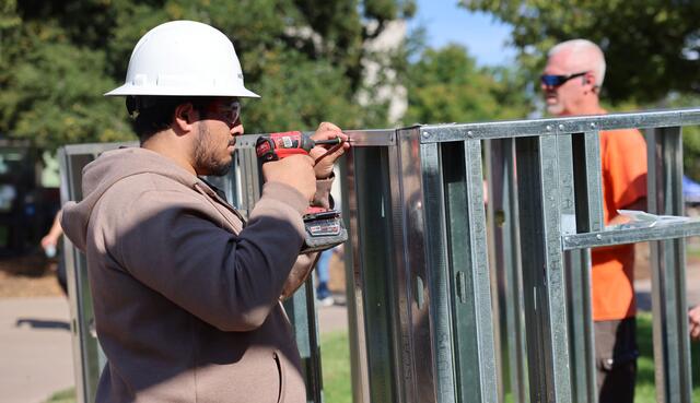 Student wearing a hard hat uses a power drill to fasten metal framing