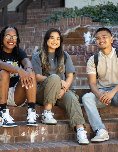 Three students sitting on steps smiling with fountain in the background