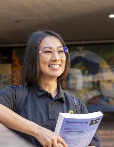 Student holding textbook leaning on bridge looking to the distance and smiling
