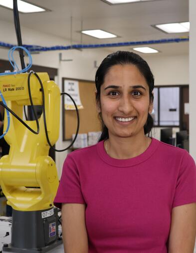 Smiling student standing in a robotics lab next to a yellow industrial robotic arm.